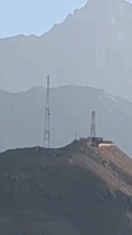 The video starts with a close-up shot of two communication towers situated on a hilltop, backed by a mountain range shrouded in mist. The camera then shifts perspective, capturing a wider landscape view from a moving vehicle. The scenery features a dry, rocky terrain with mountains in the background under a clear blue sky. A motorcyclist is visible riding along a dirt road in the distance.