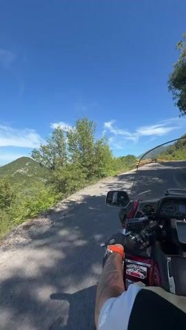 This is a first-person perspective video of someone riding a red motorcycle on a winding road. The rider's arm, wearing an orange glove, and part of their torso are visible in the foreground. The motorcycle has a windshield and mirrors. The road is paved and appears to be in a mountainous area with lush green trees and vegetation on either side. A yellow guardrail is visible along the left side of the road. The sky is bright blue with a few wispy clouds. The video captures the sensation of movement and the scenic beauty of the environment.