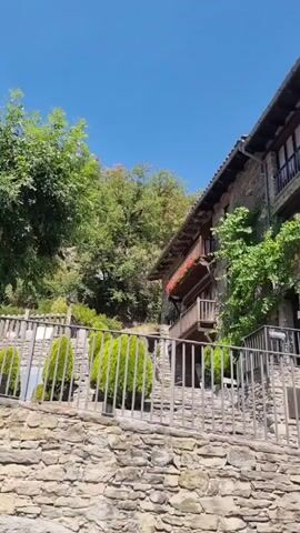 A slow pan to the right reveals a traditional stone building with a wooden balcony adorned with red flowers. The building is nestled into a hillside covered with lush green trees. In the foreground, a low stone wall with a metal fence separates the viewer from a garden area. Several manicured evergreen bushes are visible behind the fence. A large tree with full green foliage stands prominently to the left. The sky is a clear, bright blue.