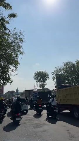 A sunny day with a bright sky and some clouds. The scene depicts a street with moderate traffic, consisting of motorcycles, cars, and a truck. Several people are riding motorcycles, and their faces are obscured by helmets or the angle of the shot. A white car is stopped in traffic. A large yellow truck is visible on the right side of the frame. Trees line the sides of the road, and some buildings can be seen in the background. The overall impression is a typical urban street scene on a clear day.