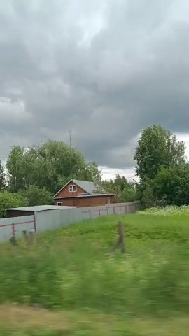 The video shows a blurred view of trees and vegetation from a moving vehicle. A small, red-sided house with a white roof is visible through the trees on the right side of the frame. There is also a large, grey structure with red markings in the foreground. The sky appears overcast.