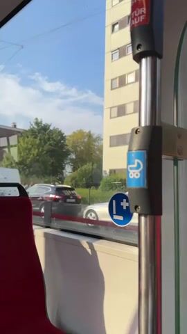 A video taken from inside a bus or tram shows a view through the window. A red seat is visible in the foreground on the left. The window looks out onto a street scene with buildings and trees. A dark colored car and a white van are driving on the road. There are also signs visible on the inside of the vehicle, one of which depicts a stroller and another a blue circle with an 'L' and a plus sign inside. The sky is blue with some clouds. A man is seen walking on the sidewalk in the distance. The sunlight is casting shadows.
