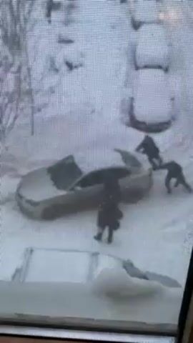 The video shows a group of four people gathered around a car that is covered in snow. The car is parked in a snowy area, and the person filming is observing from an indoor setting. The four people are seemingly inspecting or attempting to maneuver the car, possibly clearing it of snow or trying to move it, as evidenced by their positions around the vehicle and the open trunk.  The surroundings are also covered in snow, indicating a wintry environment.