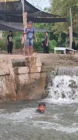 The video shows a group of children playing in a shallow waterfall area, likely a natural pool or a man-made water feature. The scene is set outdoors, with lush green foliage and bamboo visible in the background, suggesting a tropical or subtropical environment. A covered seating area with a black tarp is present, providing shade.

Several children are in the water. One boy, wearing a blue and black t-shirt and shorts, is standing on the edge of the concrete structure above the waterfall, preparing to jump. Another boy, in a black shirt, is already in the water below, looking towards the camera with a smile. A third child, wearing a red shirt and blue shorts, is seen climbing up the side of the waterfall, with water splashing around them. In the background, several adults and other children are standing or observing the activity.

The time of day appears to be daytime, with bright lighting. The weather seems to be warm, as the children are comfortably playing in the water. The atmosphere appears joyful and playful, with the children actively engaging in their water activities. There is no visible text in the video. The location context provided, Saweuk, Indonesia, aligns with the visual cues of tropical vegetation and an outdoor recreational setting.