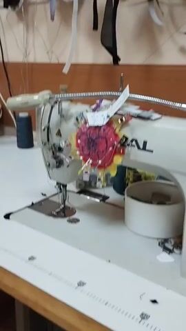 The video shows a sewing room. The focus is on a white industrial sewing machine with a colorful flower decoration attached. To the right of the sewing machine, there are shelves and drawers filled with various sewing supplies, including spools of thread, a pincushion, measuring tapes, and scissors. The shelves also hold fabric and what appear to be organized containers of items. The overall impression is of a well-equipped and organized workspace for sewing.