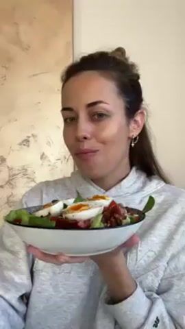 The woman holds a bowl of salad with both hands, tilting it slightly toward the camera. The salad contains lettuce, red vegetables, and two boiled eggs cut in half with runny yolks. She looks at the camera and takes a bite with a fork. She is wearing a grey hoodie and has her hair in a loose bun. Her expression is one of satisfaction while she chews, suggesting enjoyment of the meal.