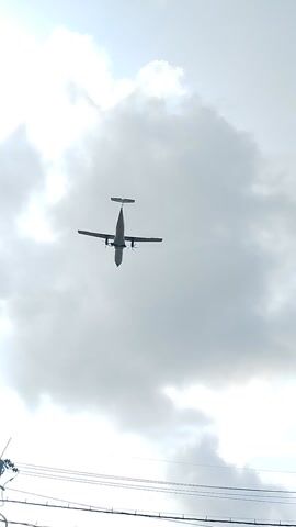 A low-angle shot captures a turboprop airplane flying against a cloudy sky. The airplane is a prominent object, appearing from a bottom-up perspective. The sky is overcast with large, white clouds dominating the frame. To the right of the airplane, utility poles and wires are visible, suggesting an urban or semi-urban setting. In the far right of the frame, part of a building with a red sign that reads "J&T Express" is visible, confirming the location as Sedati Gede, Indonesia. The lighting suggests it is daytime, possibly late afternoon due to the diffused light. No people are visible in the frame. The overall mood is neutral, with the focus on the airplane and its surroundings.