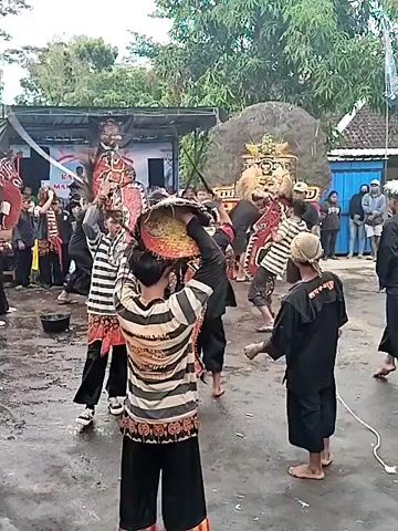 The video shows a group of people gathered outside, likely for a traditional performance or festival. A large, decorative lion head is prominently displayed, along with several individuals dressed in striped shirts, black pants, and elaborate headgear. Some of them are carrying what appear to be cultural objects, possibly props for the performance. A few people are holding flags, while others stand around observing the scene. The ground is wet, suggesting it may have recently rained. The setting appears to be a village or rural area, with houses and trees visible in the background.