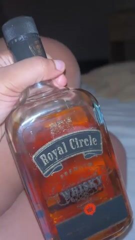 The video shows a hand holding a bottle of Royal Circle Premium Extra Smooth Whisky. The bottle is partially full and the label is clearly visible. The background is blurry but appears to be a bed.