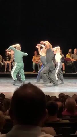 A performance is taking place on a stage in front of a seated audience. On stage, six performers, dressed in casual athletic-style clothing in shades of green, gray, and black, are dancing in a group. The performers are facing away from the camera, their arms raised and in motion. The stage is lit from above, casting shadows. The audience is visible in the foreground, with the backs of their heads and shoulders filling the bottom of the frame. The environment appears to be a theater.