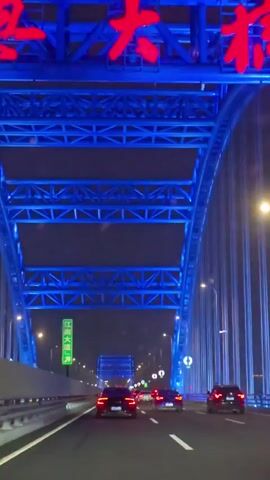The video shows a first-person perspective from inside a moving vehicle driving on a bridge at night. The bridge's structural elements are illuminated with bright blue lights, forming a distinct archway overhead. Multiple cars are visible ahead on the multi-lane road, all with their red taillights illuminated. Streetlights cast a warm yellow glow along the side of the road and on the bridge structure. A green road sign with white Chinese characters is visible on the left side of the road. The sky is dark.