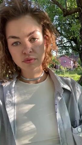 A young woman with curly, reddish-brown hair and green eyes is featured in a selfie video filmed outdoors during what appears to be the golden hour, possibly late afternoon or early evening, given the warm, directional light and visible shadows. She is wearing a light-colored t-shirt, an open light-blue button-up shirt, a sparkling choker necklace, and dangling earrings.

The video begins with her looking directly at the camera with a neutral expression. She then tilts her head, still looking at the camera. Finally, she closes her eyes, smiles, and tilts her head back, seemingly enjoying the sunlight on her face.

The background shows lush green trees and grass, indicative of a garden or rural setting. A light-colored house with a red roof is visible in the distance through the foliage. The sky is clear blue. The scene suggests a peaceful, natural environment in Danilovgrad, Montenegro.