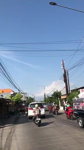 The video shows a street scene with a clear blue sky above. Several vehicles are visible, including a white SUV and a motorcycle in the foreground, with a person on the motorcycle. A red and white tower, possibly a communication tower, is prominent in the background. Overhead, a complex network of electrical wires stretches across the sky. On the sides of the street, there are buildings, some with vegetation, and what appear to be small businesses or stalls. The overall impression is of a busy street in a populated area.