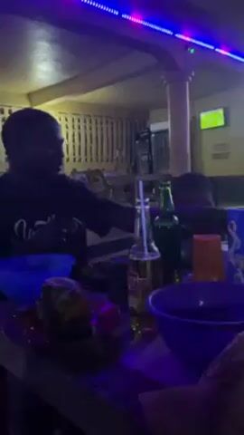 The video shows people sitting around a table in what appears to be an outdoor or semi-outdoor dining area, possibly a bar or restaurant.  There are at least three adults and one young child visible. The table is cluttered with dishes, bowls, bottles of beer, and a carton of milk.  The area is illuminated by overhead lighting, including blue LED strips.  A TV is visible in the background, possibly showing a sports game. One person at the table is wearing a shirt with the word "Hustle" on it.  One of the adults is holding a child on their lap. The overall atmosphere appears casual and relaxed.