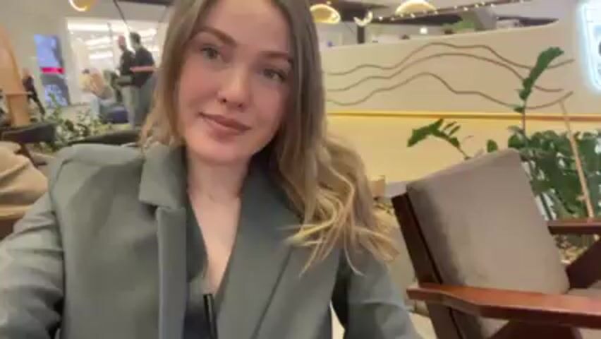 A woman is sitting in a café or restaurant in what appears to be a shopping mall. She is wearing a grey blazer and drinking from a clear plastic cup with a black straw. Her hair is blonde and shoulder-length. In the background, there are other people, plants, and store signs. The café has wooden chairs and tables, and the lighting is bright.