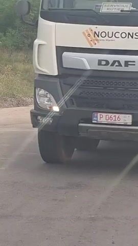 The video begins with a close-up of a white truck's front bumper and headlight. The truck has a green and white sticker with the number 6383 and "DAF" written below it. The scene then shifts to a wider view of a dusty road where two men are working. One man, wearing an orange hat and a blue t-shirt, is sweeping the road with a broom. Another man, wearing a yellow safety vest and a blue cap, is digging with a shovel. A red and yellow striped traffic cone is visible behind them. The video then shows the front of an orange SUV driving on the road. The SUV has a license plate that is partially visible, showing "27". The video returns to the men working on the road, with the man in the yellow vest continuing to dig. The environment appears to be rural, with trees and vegetation in the background.
