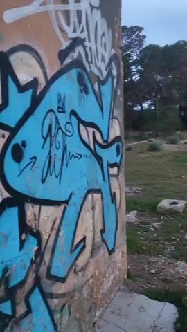 The video shows a brief view of an abandoned structure covered in graffiti. The exterior walls are painted with blue, black, and white graffiti tags. The video moves inside the dark structure revealing more graffiti on the interior walls, including green and red lettering. Debris covers the floor. Light enters through openings in the walls.