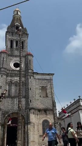 The video shows the Danza de los Voladores, a traditional Mesoamerican ceremony, being performed in front of a large stone church. The main focus is a tall pole erected in front of the church, with four men in traditional red and white clothing attached to ropes at the top. The men are performing the dance, swinging in a circular motion as they descend from the pole. One man remains at the top of the pole, playing a flute and drum, controlling the pace of the descent. The church behind them is made of gray stone and has a prominent tower with a cross at the top. A few people stand on the ground watching the performance, including a man in a blue shirt who appears to be speaking to someone. The sky is blue with scattered clouds, and the overall setting appears to be a sunny day in a public square.