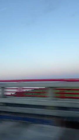 The video shows a view from a moving vehicle, likely a car, traveling over a bridge. A red railing with horizontal white bars is prominent in the foreground, with a blue stripe along the bottom edge of the railing. Beyond the railing, a body of water, possibly a river or lake, is visible under a clear sky transitioning from light blue at the top to pale pink and purple near the horizon, suggesting either sunrise or sunset. The distant landscape features green fields and trees. The movement of the vehicle causes a slight blur in the scenery. No people or specific actions are discernible.