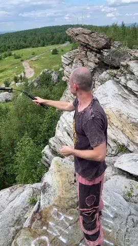 A bald man wearing a dark t-shirt and patterned pants stands on a rocky outcrop overlooking a wooded valley. He is practicing with nunchaku, swinging them in a circular motion around his body. The sky is partly cloudy.