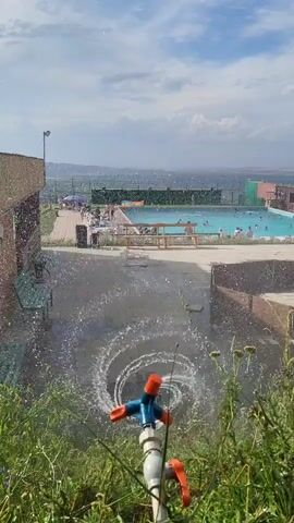 A sprinkler sprays water in a circular pattern on the ground. In the background, a swimming pool is visible, with several people swimming and lounging. There are also buildings and trees surrounding the pool. The sky is cloudy.