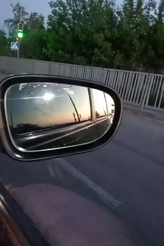 A car is driving down a road. The side mirror is visible, reflecting a sunset sky. A traffic light with a green light and a pedestrian crossing sign can be seen in the background. The car is traveling on a gray paved road next to a barrier, with trees visible beyond.