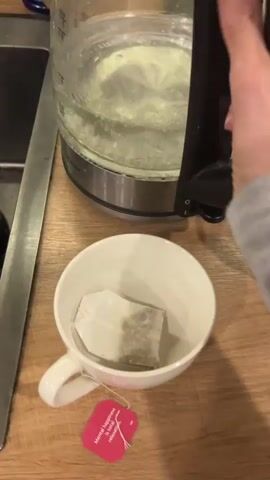 The video shows a white mug on a light brown wooden surface. Inside the mug is a tea bag with a red tag. The tag has white lettering that reads "Mental happiness is total relaxation". A stainless steel kettle is used to pour hot water into the mug. The camera angle is from slightly above and is steady. The background includes a stainless steel sink.