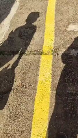 Two people's shadows are cast on a concrete platform with a yellow line down the middle. One shadow is on the left, and the other is on the right. Both shadows face away from the camera. The platform has a dirt strip to its right, which contains a set of railroad tracks. Trees are visible on both sides of the tracks. Other people are visible in the distance on the platform. The sky is bright.