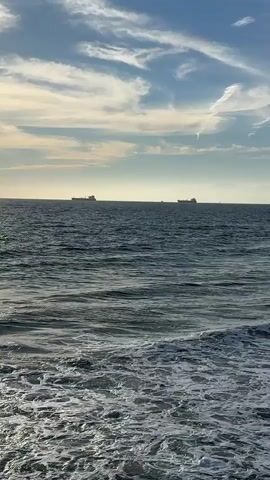 The video shows a body of water, likely the ocean, with waves crashing on a shore. In the distance, several cargo ships are visible on the horizon under a partly cloudy sky. The sun is setting, casting a warm glow on the water and clouds. The scene is peaceful and serene.