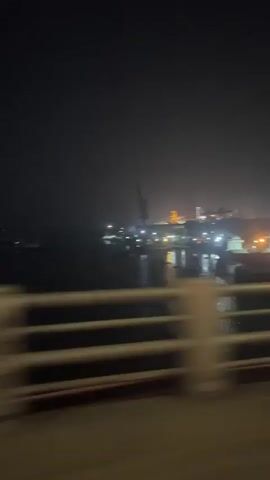The video captures a nighttime scene of an industrial waterway, likely a port or shipyard, from the perspective of someone on a moving bridge. In the foreground, a blurry bridge railing indicates motion.

The central part of the frame features a body of water reflecting numerous artificial lights from the surrounding industrial infrastructure. Several large vessels, appearing to be cargo ships or barges, are visible on the water. Towering cranes, characteristic of port operations, stand prominently in the background and on the banks, suggesting loading or unloading activities. Some of these cranes appear to be actively positioned or in operation, with lights illuminating their structures.

Beyond the water, a sprawling industrial complex stretches into the distance, distinguished by numerous bright lights illuminating buildings and structures against the dark night sky. The overall atmosphere is hazy, causing the lights to diffuse and creating a somewhat soft, glow-like effect. No specific text or identifiable landmarks are clearly visible. The scene evokes the busy, working environment of a major industrial hub after dark.