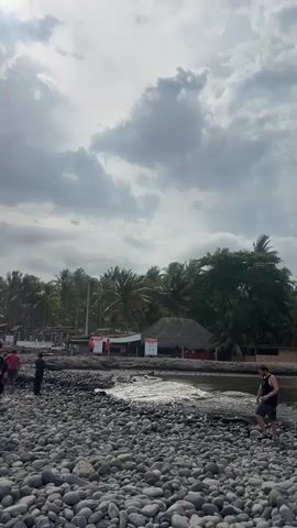 The video shows a rocky beach with several people. In the foreground, two young people stand facing away from the camera, looking towards the ocean. Behind them, a few other people are scattered on the beach, some walking and others standing near some structures. There are palm trees and buildings visible in the background. The sky is cloudy and the lighting is somewhat dim, suggesting it might be overcast or late in the day. The waves are breaking on the shore. The overall scene depicts a coastal area with some human activity.