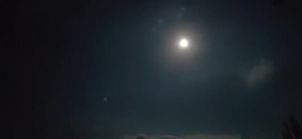 A short video captures a nighttime sky illuminated by a bright, full moon. The moon is positioned in the upper right quadrant of the frame, casting a glow that creates a soft halo effect. A faint cloud formation is visible to the right of the moon. In the lower portion of the sky, along the horizon, hints of dark shapes that could be trees or structures are present, partially obscured by what appear to be wispy clouds or fog. To the left of the moon, a smaller, fainter light is visible. The overall impression is of a clear night sky with a prominent moon.