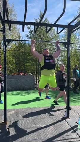 The video shows a man wearing a black weighted vest, neon yellow athletic shorts, and neon yellow sneakers doing pull-ups on a metal bar. The bar is part of a larger outdoor workout structure with "YOUSTEEL" printed on its supports. The workout takes place on a green turf and black tile surface, surrounded by trees on a sunny day. Several other people are standing around watching and encouraging the man, including one in a similar vest and another wearing a red cap.
