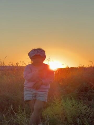 A young girl is standing in a grassy field with tall dry grass and brush. She is wearing a pink sweatshirt with the letter M on it, light blue and white striped shorts, and a patterned headband. The sun is setting behind her, casting a warm orange glow across the sky and illuminating the field. The girl is looking at the camera with a serious expression and has her arms bent at the elbows with her hands near her chest.