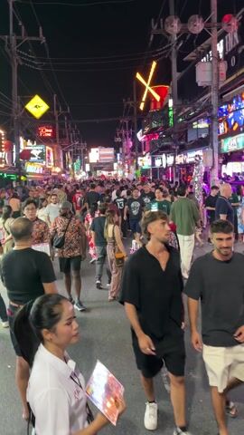 The video shows a crowded street at night, full of pedestrians. There are many shops and businesses, their bright neon signs illuminating the scene. Most people are dressed casually and walking in various directions. The street is filled with signs, utility poles and wires, and advertisements, adding to the hustle and bustle. The general environment is lively, suggestive of a popular nightlife destination.