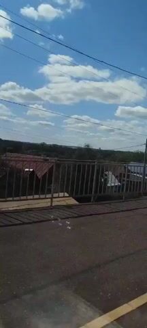 A video taken from a moving vehicle shows a daytime scene with a blue sky and white clouds. The perspective is from a road or path overlooking a rural or suburban area. In the foreground, there is a paved surface with some debris and shadows. A metal railing and a bench are visible on the left side of the frame. Beyond the railing, there are houses with varied roofs, trees, and utility poles with wires. The vehicle appears to be moving forward, causing a slight blur and a sense of motion. The scene suggests a journey through a developed landscape.