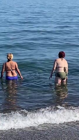 Two women are standing in the shallow water of the sea, facing away from the camera. The woman on the left has blonde hair and is wearing a blue bikini bottom and a black bikini top. The woman on the right has dark red hair and is wearing a green bikini bottom and a black bikini top. The water is clear and a gentle wave is rolling in. The background shows the vast expanse of the blue sea under a clear sky, suggesting a sunny day. The scene is likely a beach in Marbella, Spain, given the context. The overall mood appears relaxed and enjoyable, as the women are casually enjoying the water. There is no visible text in the media file.