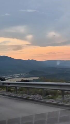 The video shows a view from a car driving on a road overlooking a valley of rolling hills and mountains. The sun is setting, casting a warm, orange glow across the sky, which is partly cloudy with dramatic grey and white formations. The landscape appears to be rural, with the hills covered in dark green foliage. In the foreground, a metal guardrail runs along the side of the road, with a paved surface visible below. There are no people or specific objects clearly identifiable beyond the natural scenery and the roadside infrastructure. The time of day is clearly sunset, and the weather appears to be clear but with significant cloud cover. The location cues suggest a scenic overlook in a mountainous or hilly region. The overall mood is serene and picturesque, capturing the beauty of a sunset in nature. No text is visible in the media file.