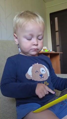 A young boy with blond hair sits on a couch, wearing a navy blue long-sleeved shirt with a cartoon dog graphic and blue shorts. He is holding a yellow and blue book open and looking down at it intently, with his finger touching the screen or page. The background shows a room with a wooden table, a door, and part of another couch. The boy appears engaged with the book, and the lighting suggests an indoor setting during the daytime.