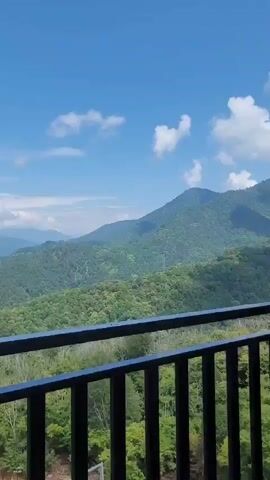 The video shows a panoramic view of lush green mountains under a clear blue sky with scattered white clouds. The foreground features a black railing, suggesting the perspective is from a balcony or a similar elevated vantage point. In the distance, houses and small structures are visible nestled within the valleys between the mountains. The overall scene depicts a serene and natural landscape.