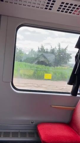 The video is shot from inside a train car, looking out a window. The window is large and rectangular, with a gray frame. Outside the window, a rural landscape is visible. There are green fields of crops, possibly corn, stretching into the distance. A dirt road runs alongside the fields, and a few buildings can be seen in the background. The sky is partly cloudy, with patches of blue sky visible between the white clouds. The train car interior is visible in the foreground. There is a red seat in the lower right corner of the frame, and a gray wall with a ventilation grate above it. A black jacket is hanging on the right side of the window. The train appears to be moving, as the landscape outside the window is blurred, indicating motion.