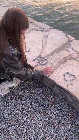 A young woman with long, straight, reddish-brown hair is seen kneeling or crouching on a pebbled shoreline next to a body of water. She is wearing a dark, possibly leather or faux leather, jacket and dark trousers, with white sneakers visible on her feet. She is actively picking up small, smooth, grey pebbles from the ground with her right hand.

On the light-colored stone surface directly in front of her, which appears to be a paved edge or wall alongside the water, she is arranging these pebbles. A heart shape made of pebbles is already visible on the stone surface to her right. To her left, she is in the process of forming another, larger, more intricate shape or design with the pebbles, which resembles letters or a symbol. Her gaze is directed downwards, focused intently on her activity.

The setting is outdoors, at what appears to be the edge of a calm lake or sea, indicated by the expanse of water in the background. The water shows subtle reflections of the sky, suggesting either early morning or late afternoon/early evening light. The ground where she kneels is covered entirely in small, rounded grey and brown pebbles. The stone surface she is working on consists of large, irregularly shaped flagstones. The overall atmosphere seems peaceful and serene, with the woman engaged in a quiet, creative pursuit by the water's edge.