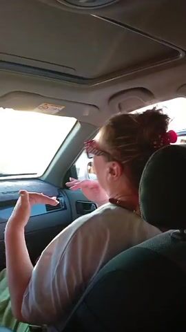 The video is a rear-facing, medium shot of a person with dark hair pulled up in a bun, wearing sunglasses and a light-colored t-shirt, sitting in the passenger seat of a car. The sun is shining brightly through the windshield, creating lens flare. The person is looking out the window and their right hand is visible near their face, appearing to gesture. The car's interior is visible, including the dashboard and part of the seats. The scene is likely outdoors during the daytime.