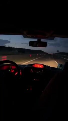 The video shows a car driving on a highway at night. The dashboard is visible, with the speedometer and radio display illuminated in red. The road ahead is lit by the car's headlights, and there are other cars in the distance. The sky is dark, with some clouds visible. The scene appears calm and focused on the act of driving.
