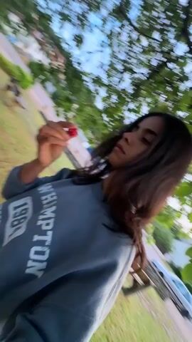A young woman with dark hair is standing outside, partially obscured by tree leaves, and eating a cherry. She is wearing a grey sweatshirt with white lettering. In the background, there is a park bench, a stone wall, a grassy area, and some buildings. The camera movement suggests the video was taken by someone walking or moving around the woman.