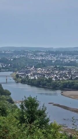 The video pans over a wide, panoramic view of a landscape with a river in the foreground and a town or city in the background. The river has a visible shoreline, suggesting it may be at a lower water level. There are trees and vegetation along the riverbanks and in the middle ground. The background features a densely populated area with numerous buildings, some of which appear to be houses with red roofs. Further in the distance, hills or mountains are visible, and a tall structure, possibly a communication tower, is present on one of the hills. The sky is overcast, with a muted, grayish tone. The overall impression is of a semi-urban or suburban area situated along a river, with natural landscapes in the surrounding area.
