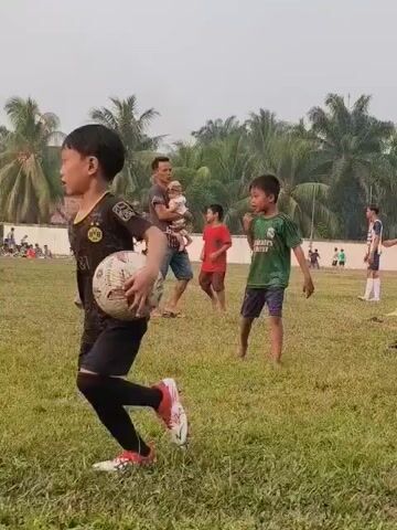 The video shows a group of children playing soccer on a grassy field. In the foreground, a young boy in a green shirt and blue shorts runs towards the right side of the frame, falling to the ground as if he tripped. A man holding a baby stands in the middle of the frame, watching the children. Other children in red and black shirts are also visible on the field. In the background, there are palm trees and a white wall. Some people are standing near the wall, and a few more children are playing soccer in the distance. The sky is overcast.
