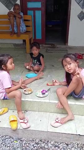 Three young children are eating food on the porch of a house. Two of the children are sitting on the steps, while the third child is sitting on the porch floor. Each child has a plate of food and a utensil. An elderly woman is sitting on a bench behind the children. The porch is tiled and there is a pile of gravel in front of it. The house has a wooden door and window frames.