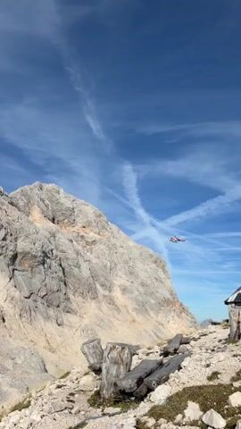 The video shows a rocky mountain range under a blue sky with wispy clouds. In the foreground, there are weathered wooden logs and rocks. A helicopter is visible in the middle distance, flying near a steep, rocky mountainside. The helicopter appears to be red and white.