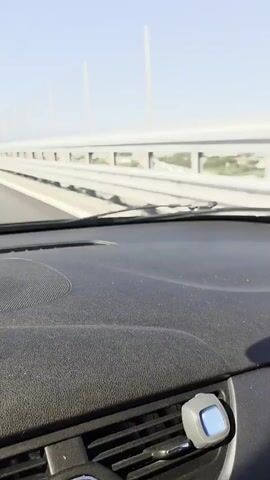 A first-person, dashcam-like video captures a view from inside a car driving on a multi-lane highway under a clear sky. The highway appears to be an elevated structure, with a white guardrail and a tall fence running along the right side, separating the road from what appears to be a lower landscape. Another car is visible in the distance, driving in the same direction. The dashboard and windshield wipers are in the foreground. A bright, sunny day is indicated by the lighting.